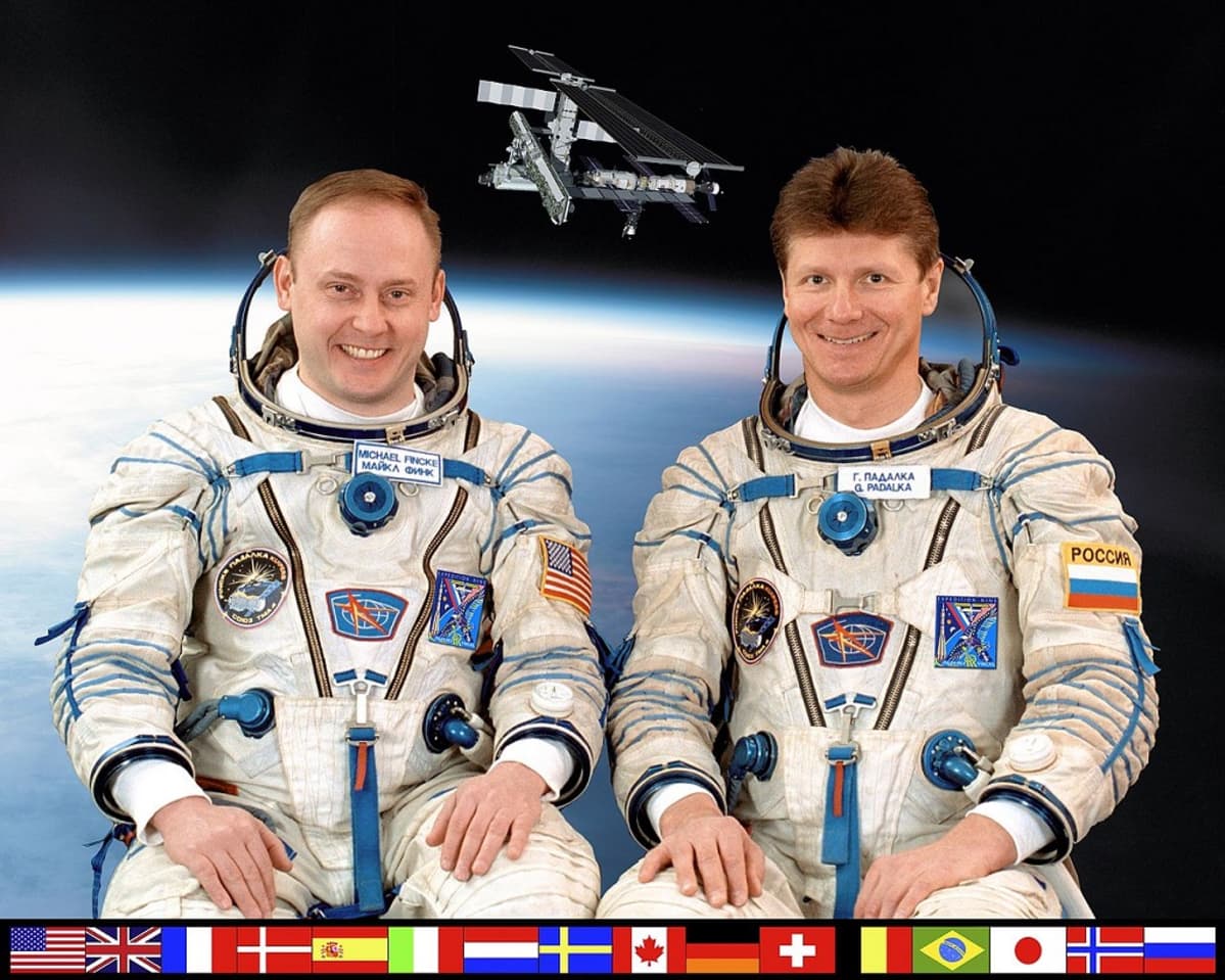 Kaksi avaruuspukuista miestä, taustalla kuva avaruusasemasta, alalaidassa 16 maan liput kertomassa, mistä maista ISS:llä on ollut väkeä.