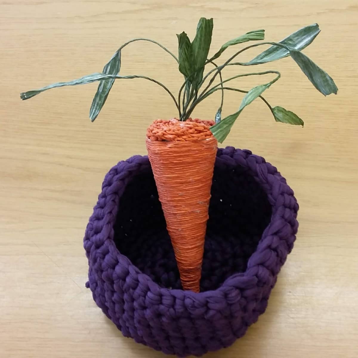 Oranssi, paperinarusta valmistettu porkkana, jossa vihreät paperiset naatit. Porkkana on violetissa virkatussa korissa.