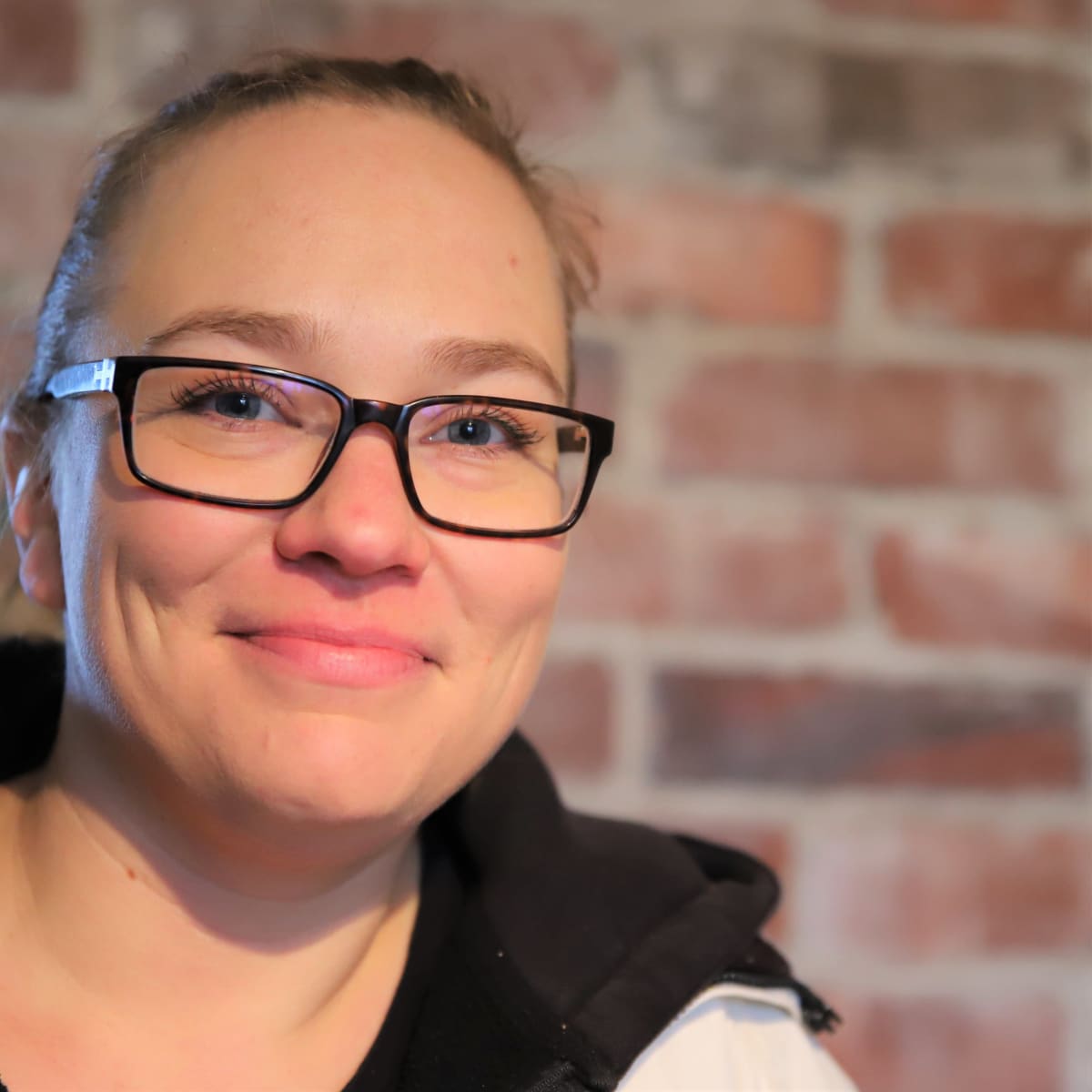 Riikka Mäkelä katsoo kameraa, taustalla vanha tiiliseinä.