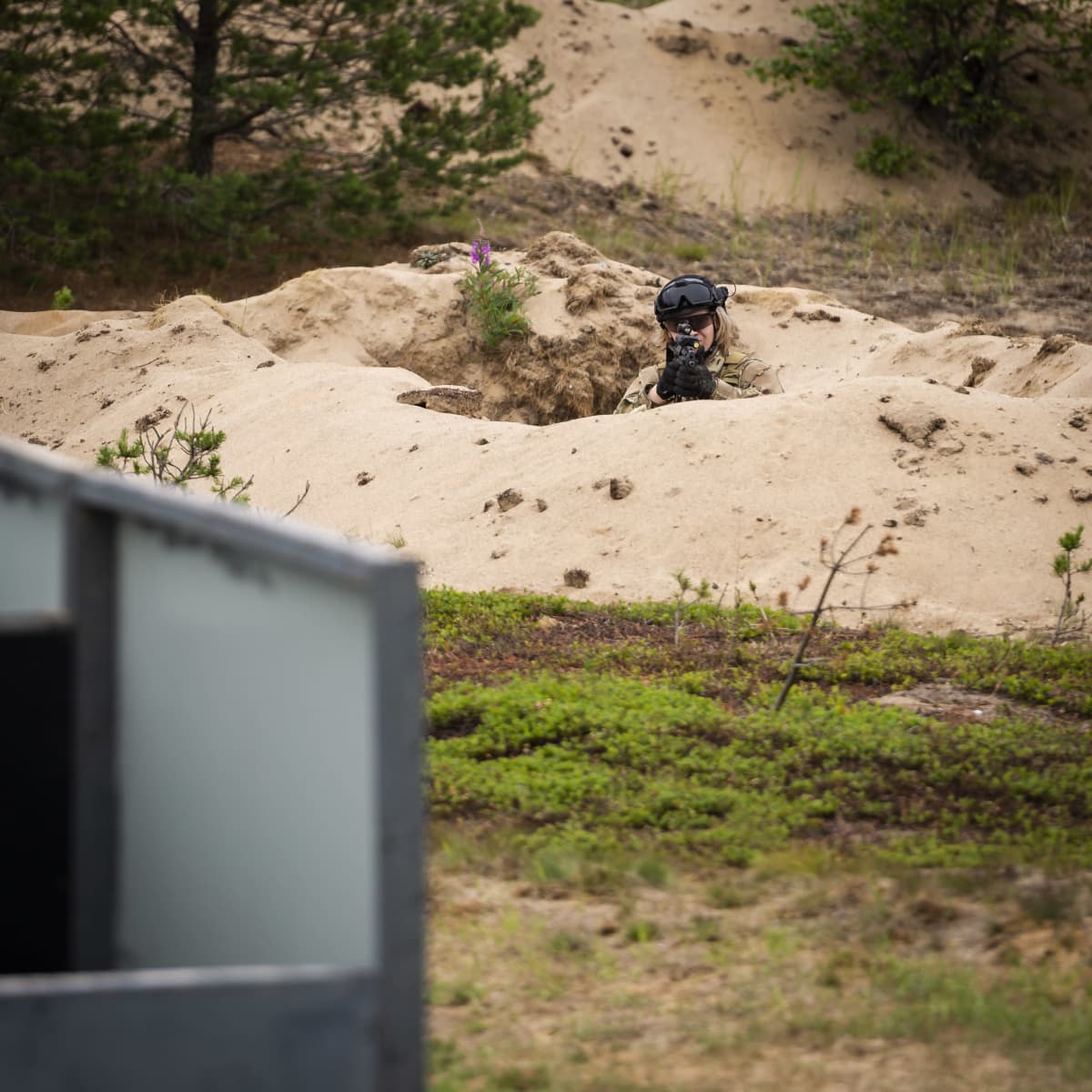 Vasemmalla alakulmassa näkyy osa Airsoft-radan rakennusta. Oikealla yläkulmassa Airsoft-pelaaja kurkistaa juoksuhaudasta ja tähtää aseella.