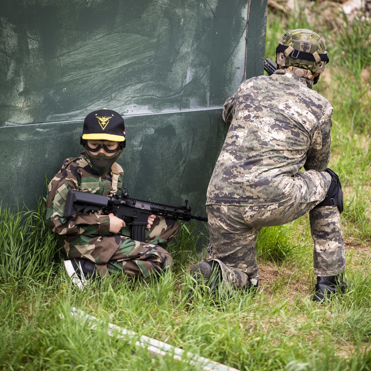 Vasemmalla nuori Airsoft-pelaaja kyyristyneenä maassa. Oikealla aikuinen Airsoft-pelaaja kurkistaa nurkan taakse.