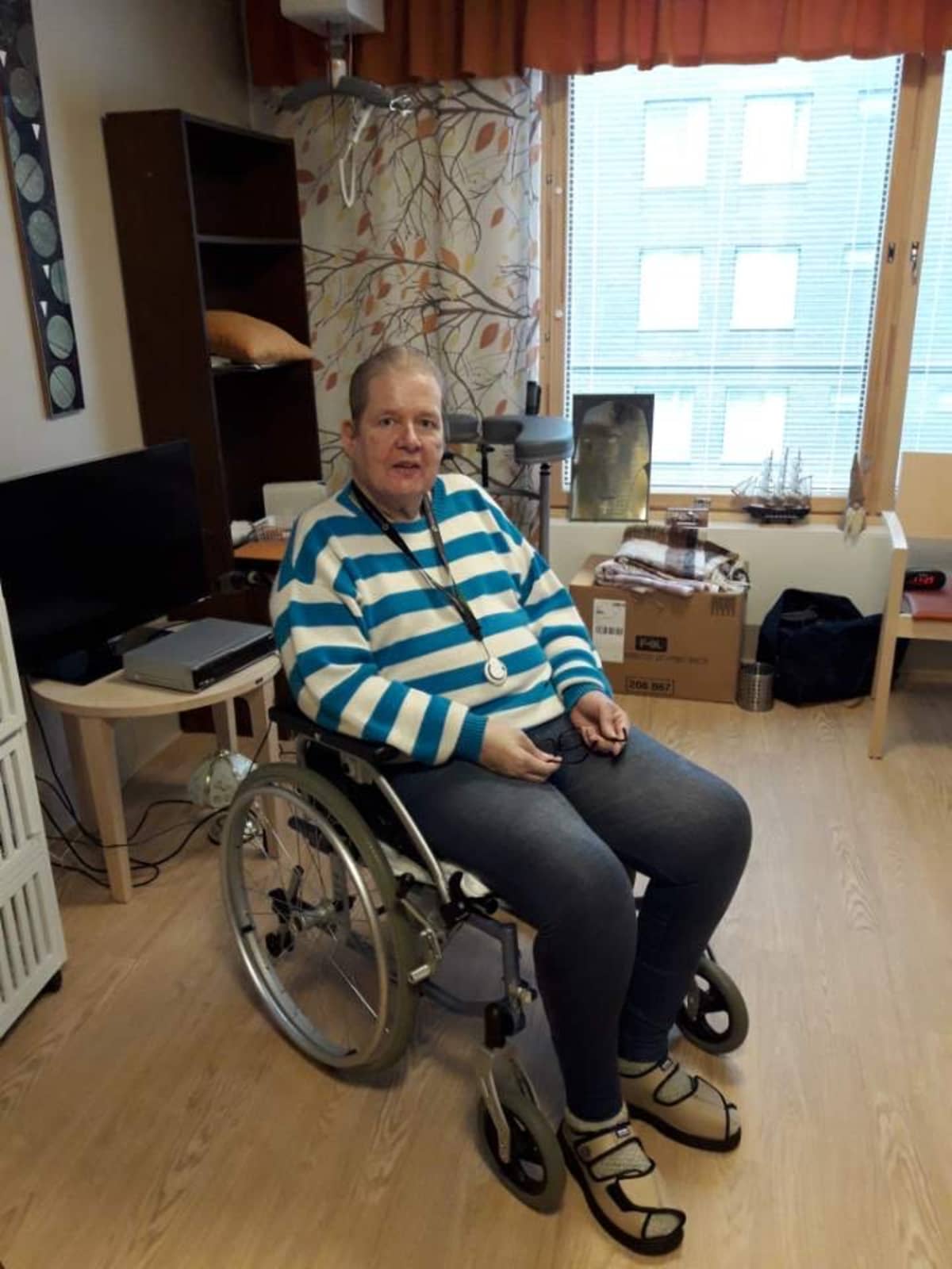 Pekka Reinikainen istuu sinivalkoraidallinen collegepaita päällään pyörätuolissa keskellä omaa huonettaan vakava ilme kasvoillaan.