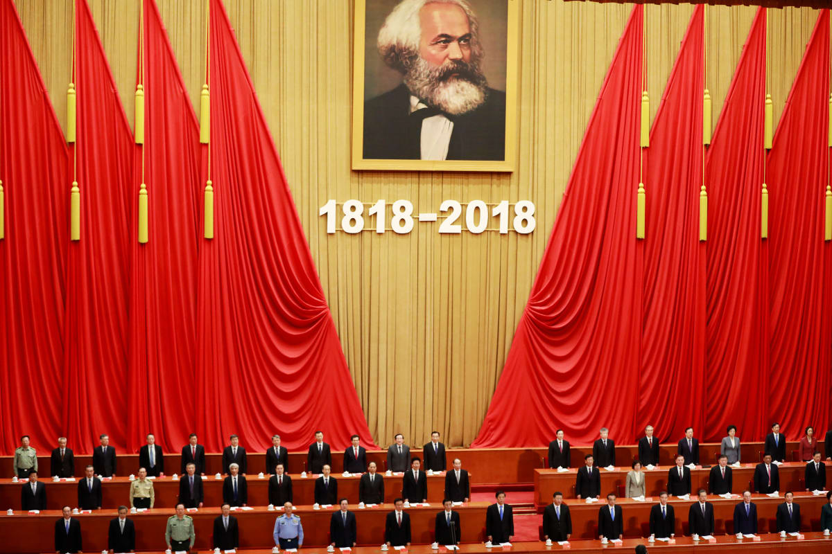 Seinällä suurten punaisten lippujen reunustamana on Marxin kuva, jonka alla lukee 1818 - 2018. Pulpettiriveillä kuvan alla seisoo joukko puoluetyöläisiä, pääosin mustissa puvuissa.