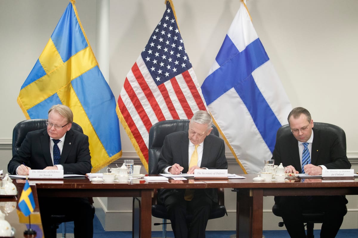 Kolmikko allekirjoittaa pöydän ääressä. Taustalla maiden liput.