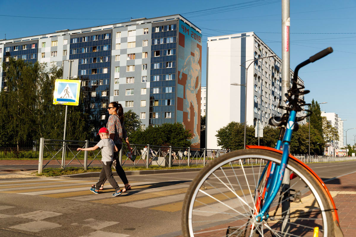 Kuvan etualalle on parkkeerattu polkupyörä. Nainen ja poika ylittävät suojatietä. Taustalla on rykelmä kerrostaloja. Yhden talon seinässä on jalkapallon MM-kisoja mainostava suuri seinämaalaus.