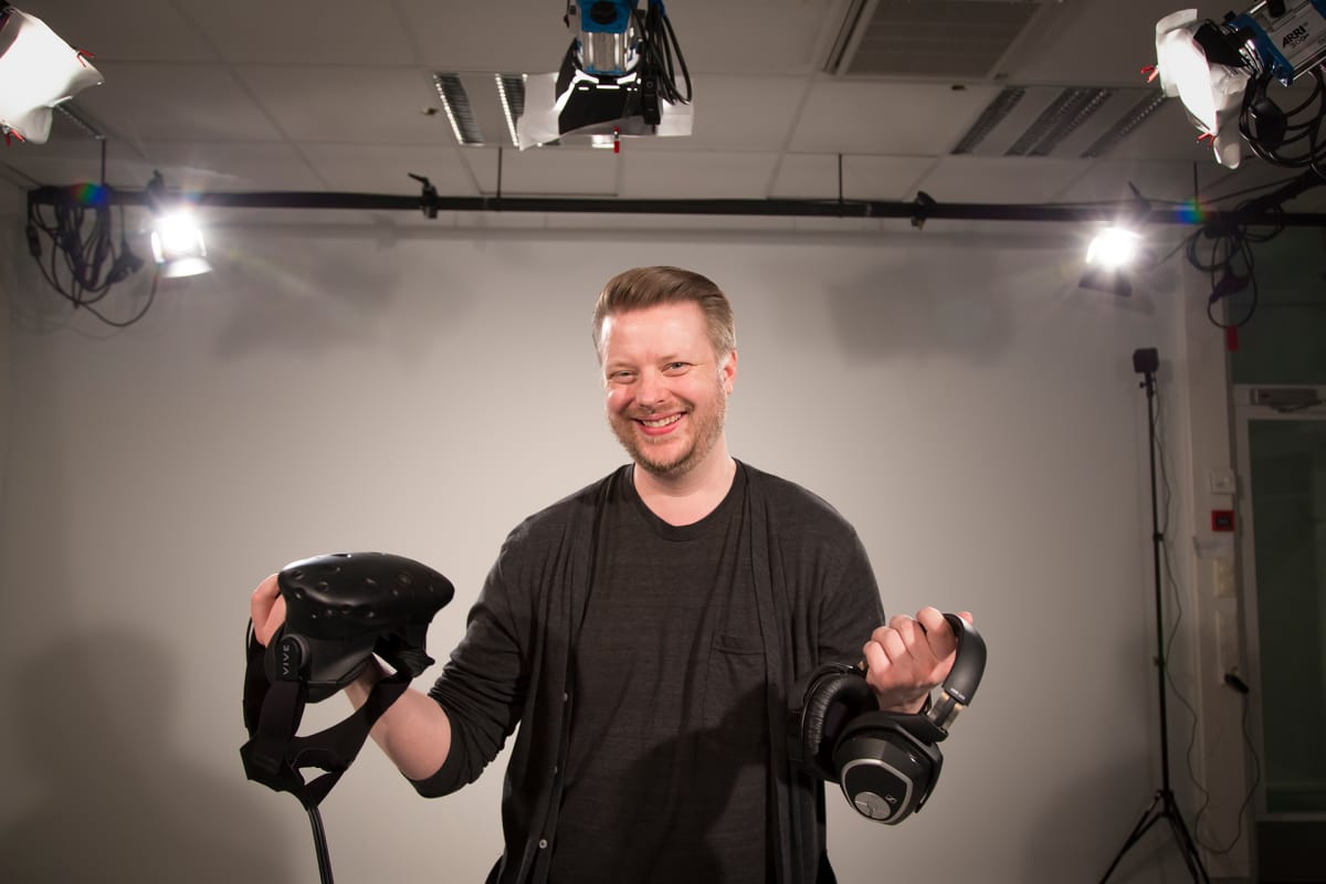 Mies poseeraa studiossa virtuaalilasit ja kuulokkeet kädessä.