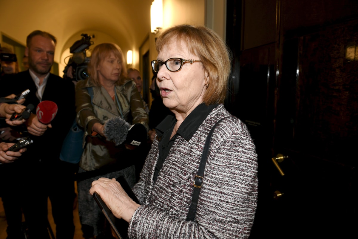 En kvinna i spräcklig kavaj utanför en stor trädörr med en skara journalister kring sig.