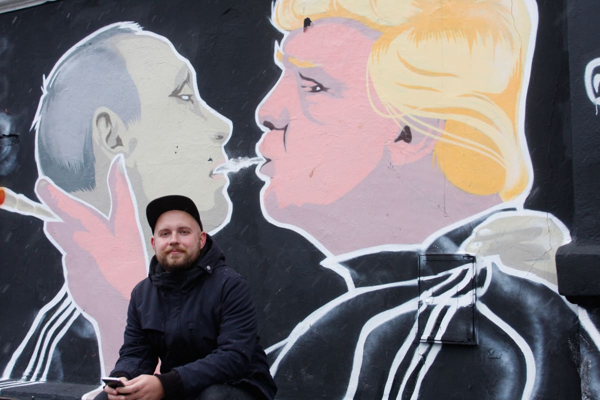Mies istuu seinämaalauksen edessä. Siinä Putinin ja Trumpin kaltaiset hahmot kaulailevat.