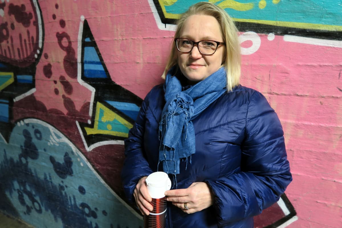 Vaaleahiuksinen nainen termari kädessä graffitiseinän edessä.