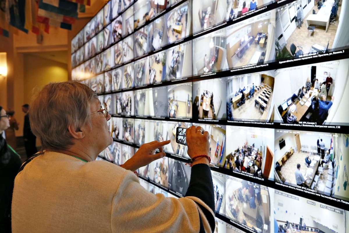 Nainen ottaa kuvia puhelimellaan, kun videonäytöt välittävät live-kuvaa Venäjän vaalien äänestyspaikoista sunnuntaina 18. maaliskuuta.