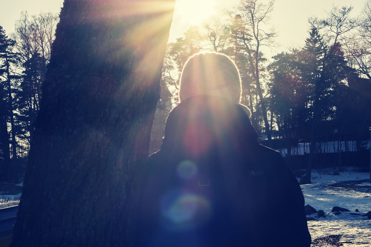 Nainen nojaa puuhun ja katselee kohti aurinkoa.