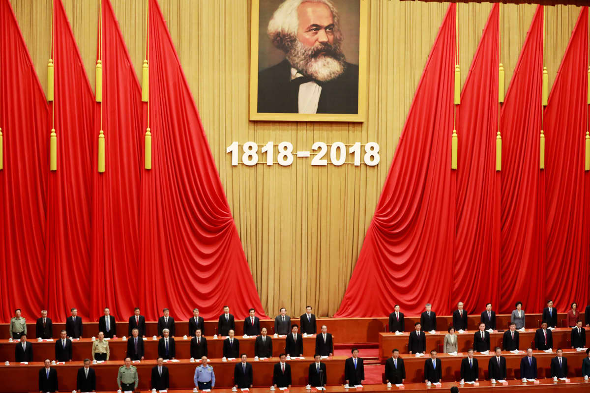 Seinällä suurten punaisten lippujen reunustamana on Marxin kuva, jonka alla lukee 1818 - 2018. Pulpettiriveillä kuvan alla seisoo joukko puoluetyöläisiä, pääosin mustissa puvuissa.
