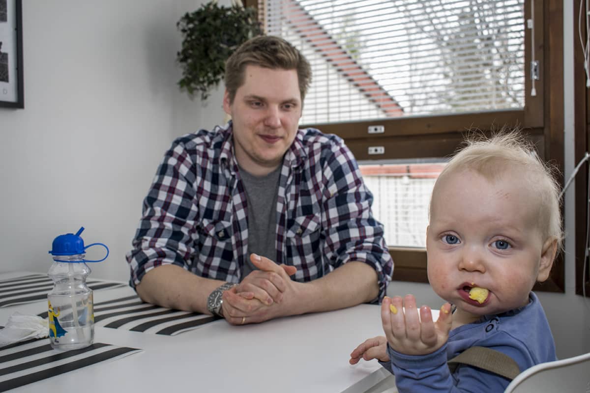 Yksivuotias lapsi istuu syöttötuolissa ja isä on vieressä