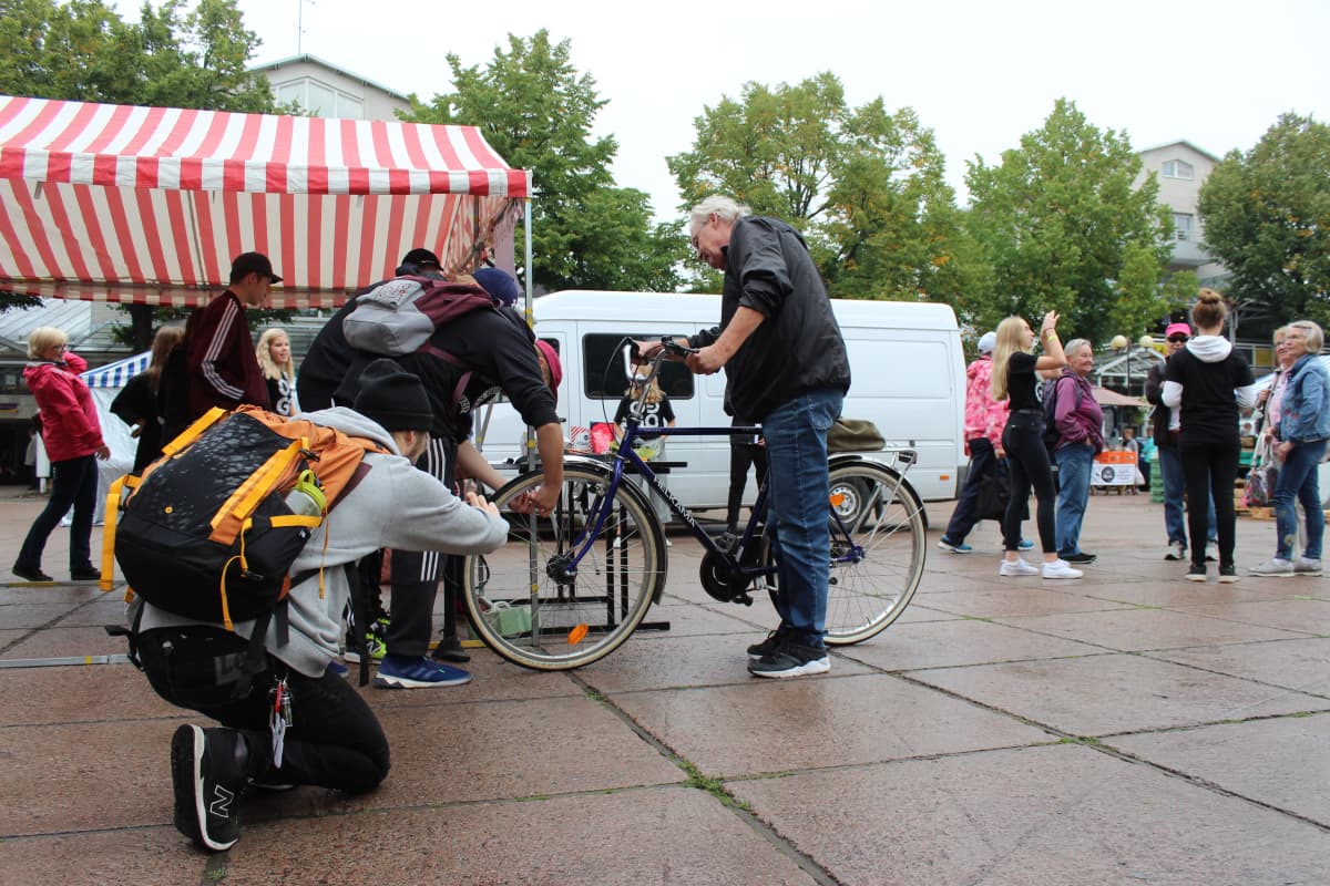 Nuoria miehiä pumppaamassa ilmaa vanhemman miehen polkupyörän etukumiin Karhulan torilla.