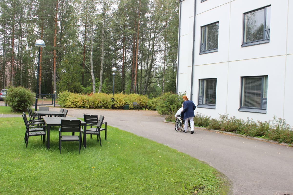 Hoitaja työntää asukasta hoivakodin pihalla pyörätuolissa.
