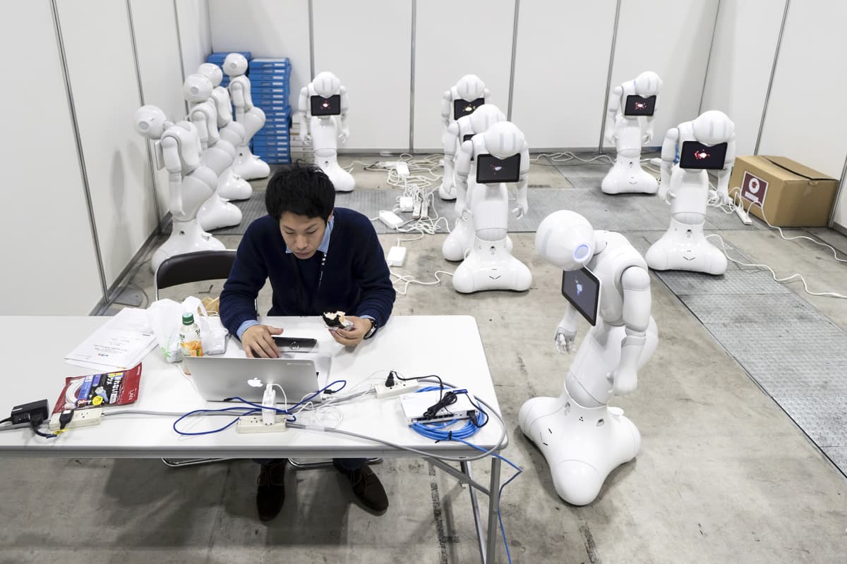 Työntekijä syö lounasta työpöydän luona läppärin edessä ja taustalla Pepper robotit latautuvat.