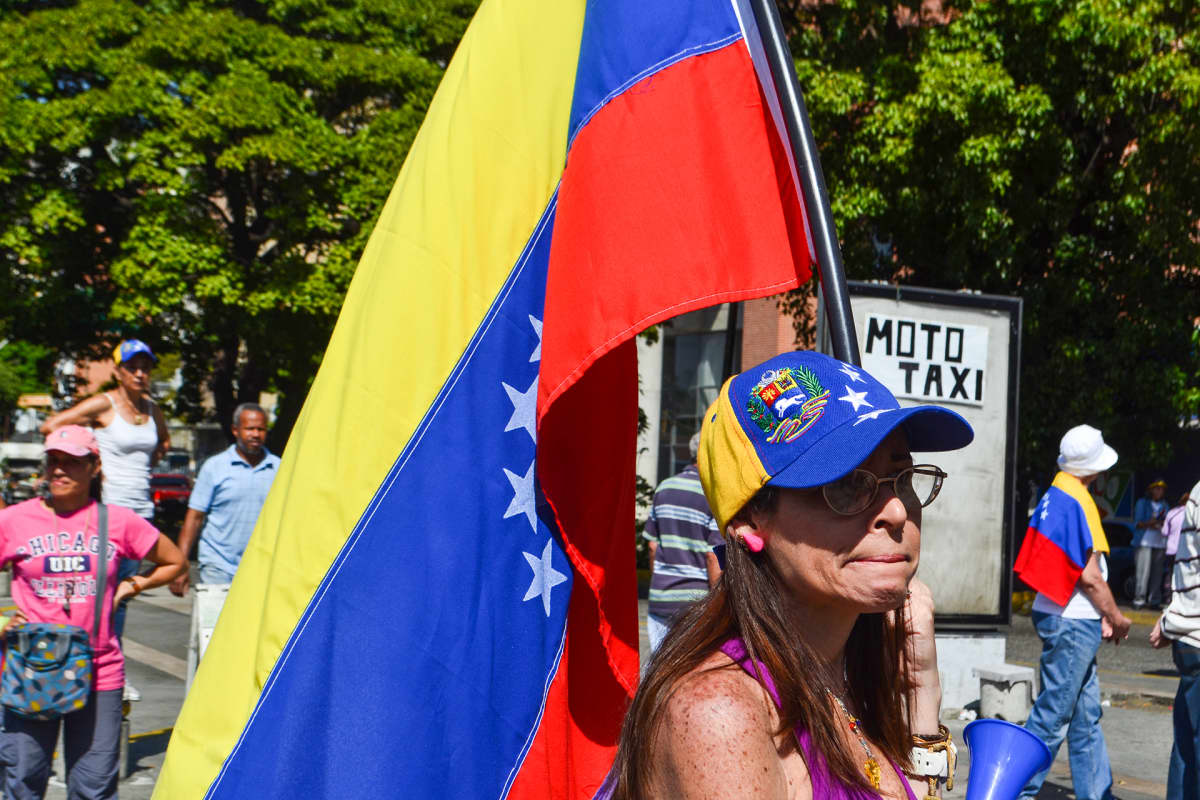 Lippalakkipäinen nainen kantaa Venezuelan lippua. Taustalla näkyy puustoa.