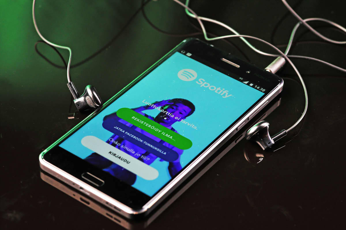 Älykännykkä ja nappikuulokkeet pöydällä. Kännykän näytöllä näkyy Spotifyn sovellus.