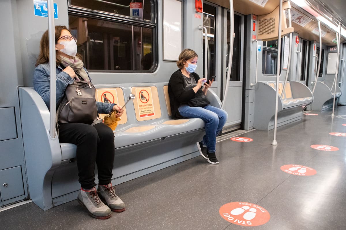 Matkustajat istuvat metrossa turvavälin päässä, turvaväliä osoittavia merkkejä Milanon metron lattiassa ja penkeillä