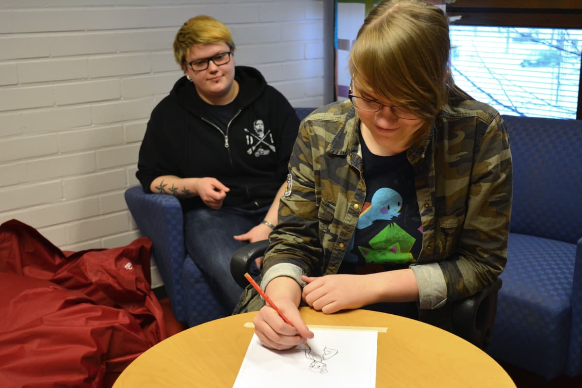 Helmi Kinnunen piirtää kuvaa naisesta, Anu Salonen katsoo taustalla Helmi Kinnusen piirtämistä