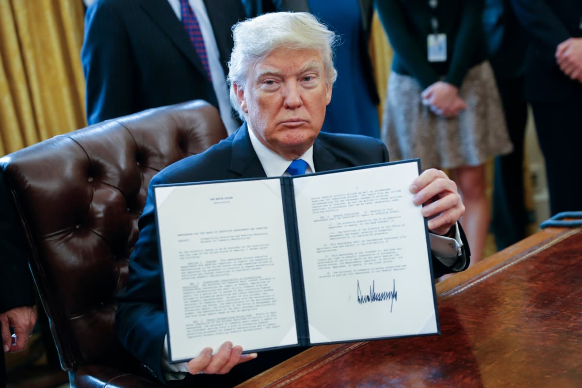 Kirjoituspöydän ääressä istuva Trump pitää käsissään ohutta kansiota, jossa on allekirjoitettu paperi.