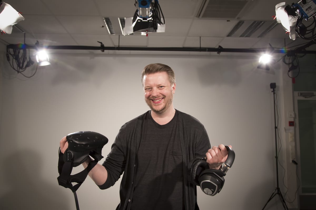Mies poseeraa studiossa virtuaalilasit ja kuulokkeet kädessä.