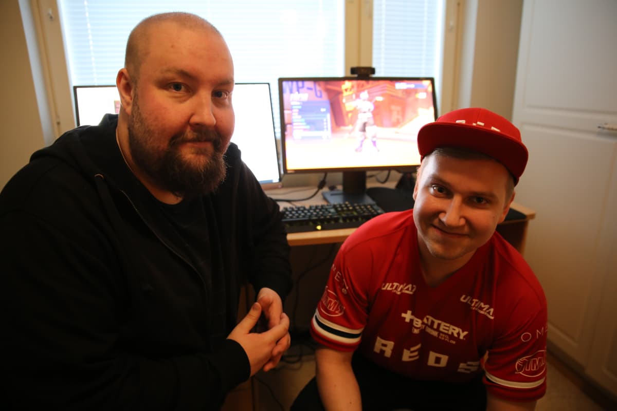 Kaljupäinen parrakas mies sekä lippalakkipäinen nuori mies poseeraa tietokoneen äärellä.