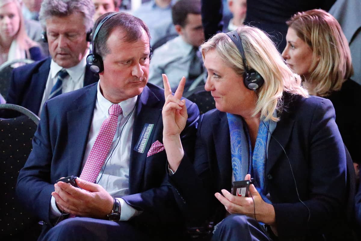 Louis Alliot ja Marine Le Pen istuvat kokoontumisessa. Molemmilla on päässään kuulokkeet. Le Pen näyttää juuri Aliot'lle oikean kätensä etu- ja keskisormea.