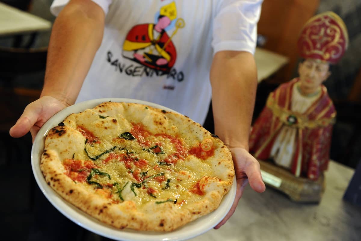 Mies ojentaa lautasta, jolla pizza, vieressään Napolin suojeluspyhimystä San Gennaroa esittävä pysti.