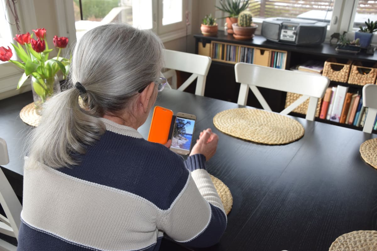 En kvinna sitter vid ett köksbord med ryggen mot fotografen. Hon tittar på ett fotografi i sin mobiltelefon