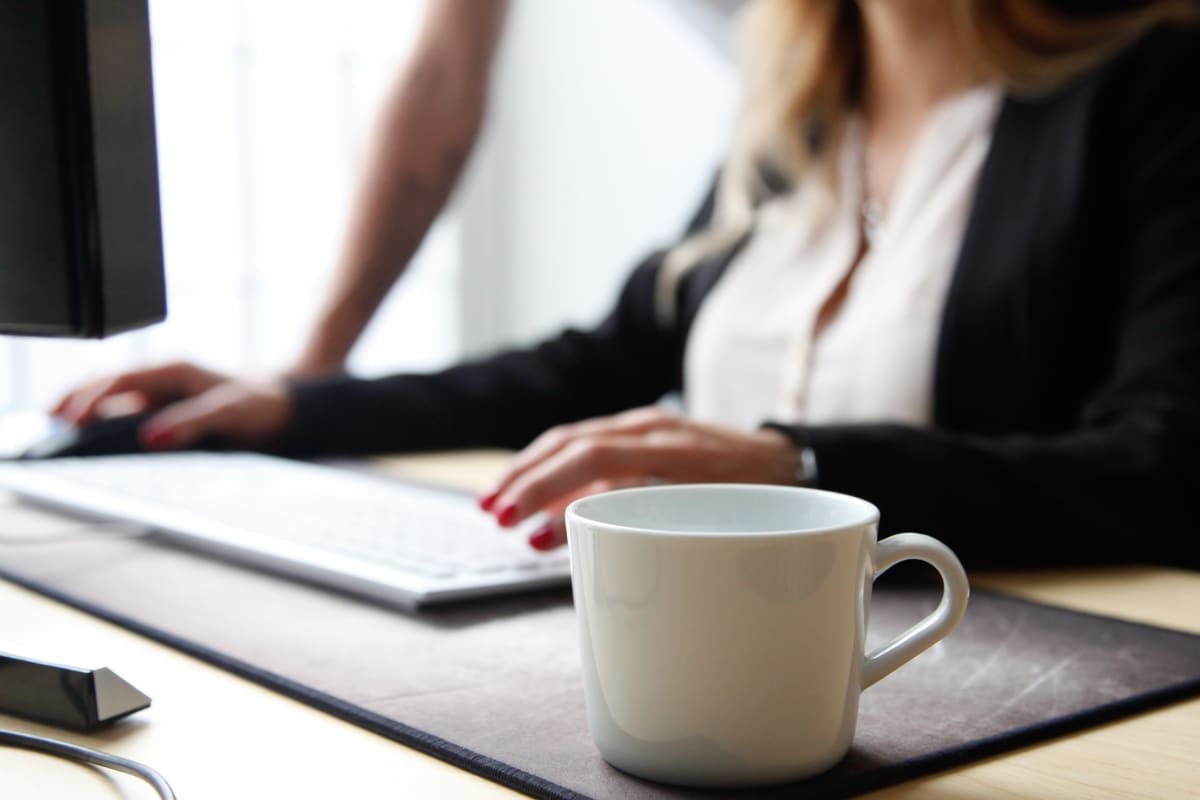 Kaffekopp i förgrunden, människa som skriver på dator i bakgrunden