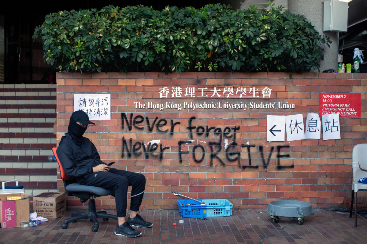 Naamioitunut opiskelija istuu seinäkirjoituksen vieressä. Englanninkielinen teksti on Never forget, never forgive.