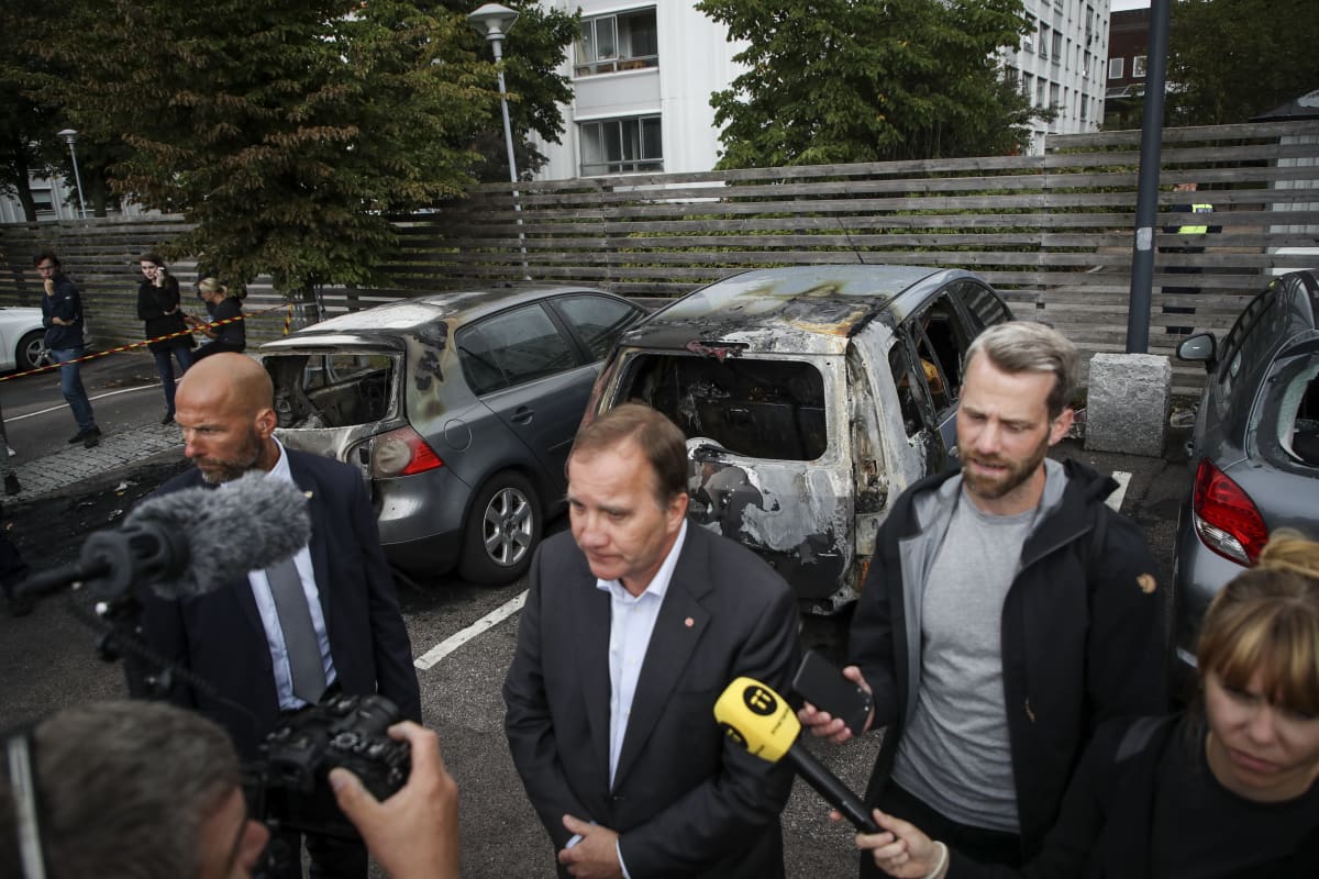 Stefan Löfven taustallaan poltettuja autoja.