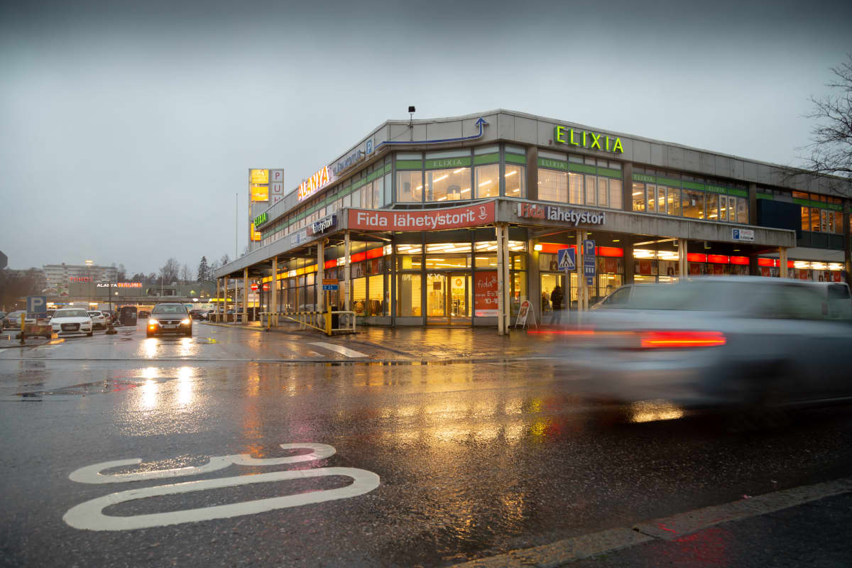 Kauppakeskus Puhos ulkoa päin. Eri liikkeiden valot loistavat sisältä lämpimästi sateisena päivänä. Liikeiden logoja: Fida, Elixia. Auto hurahtaa juuri ohi, parkkipaikalta tulevan auton valot heijastuvat märästä asfaltista.