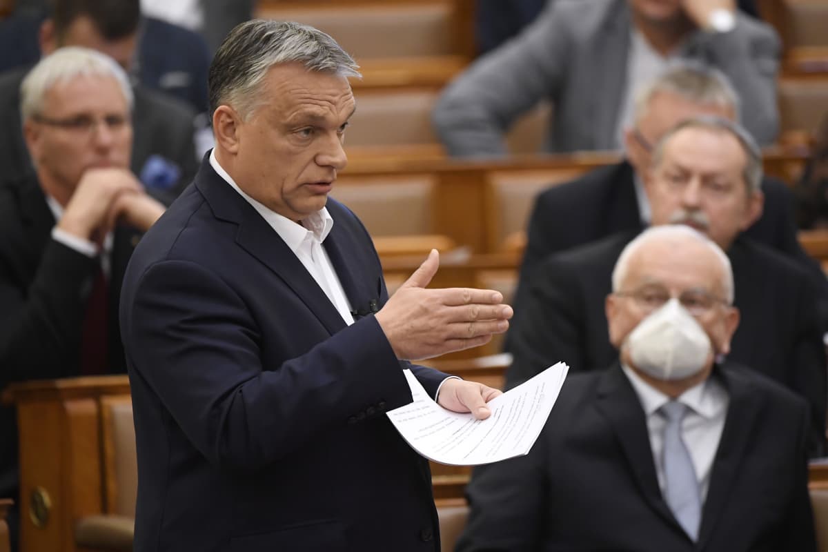 Viktor Orban puhuu paperit kädessään. Taustalla mies hengityssuojuksessa.
