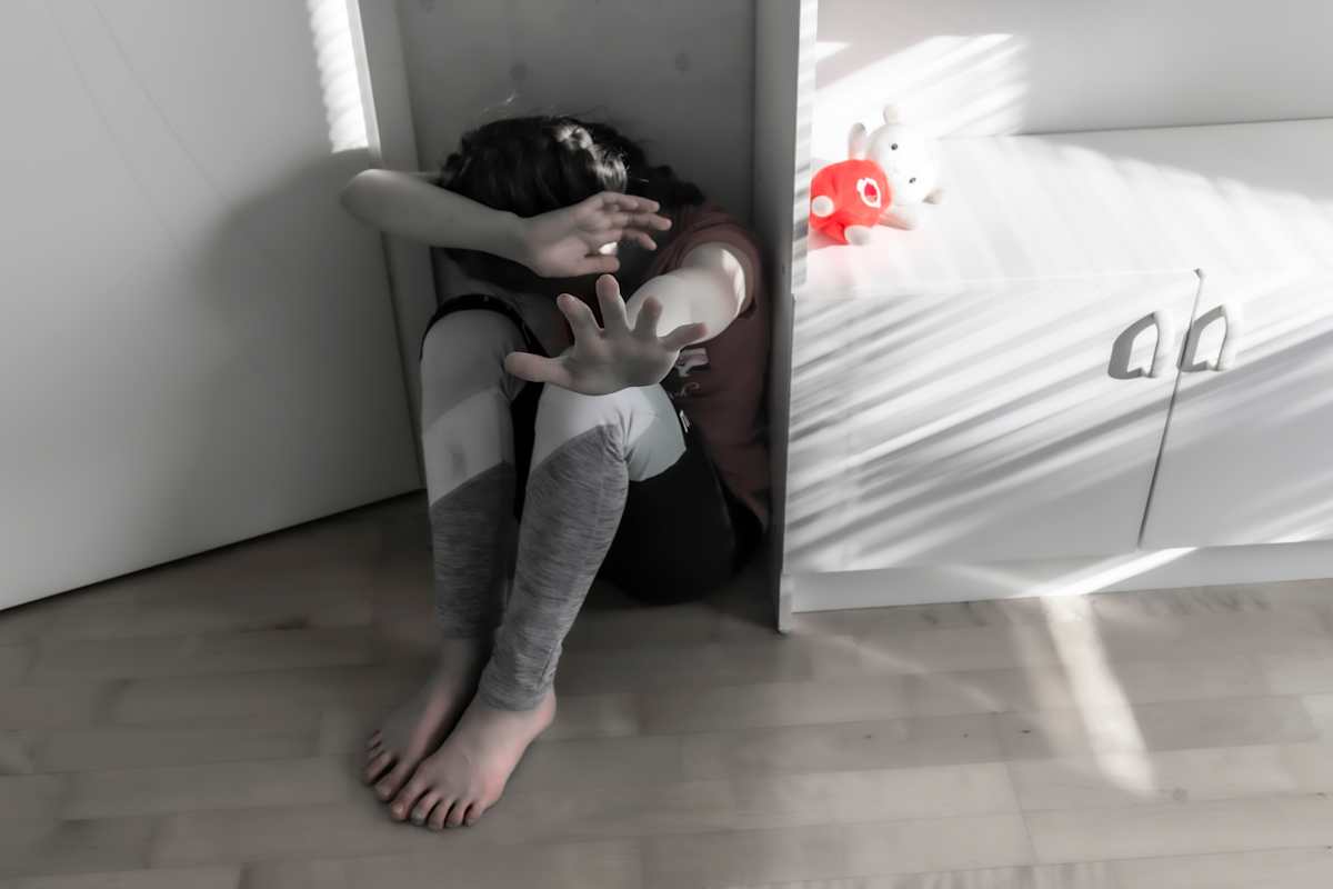 Dramatisoitu kuva, jossa lapsi istuu nurkassa suojaten kädellä päätään ja torjuen uhkaa toisella.