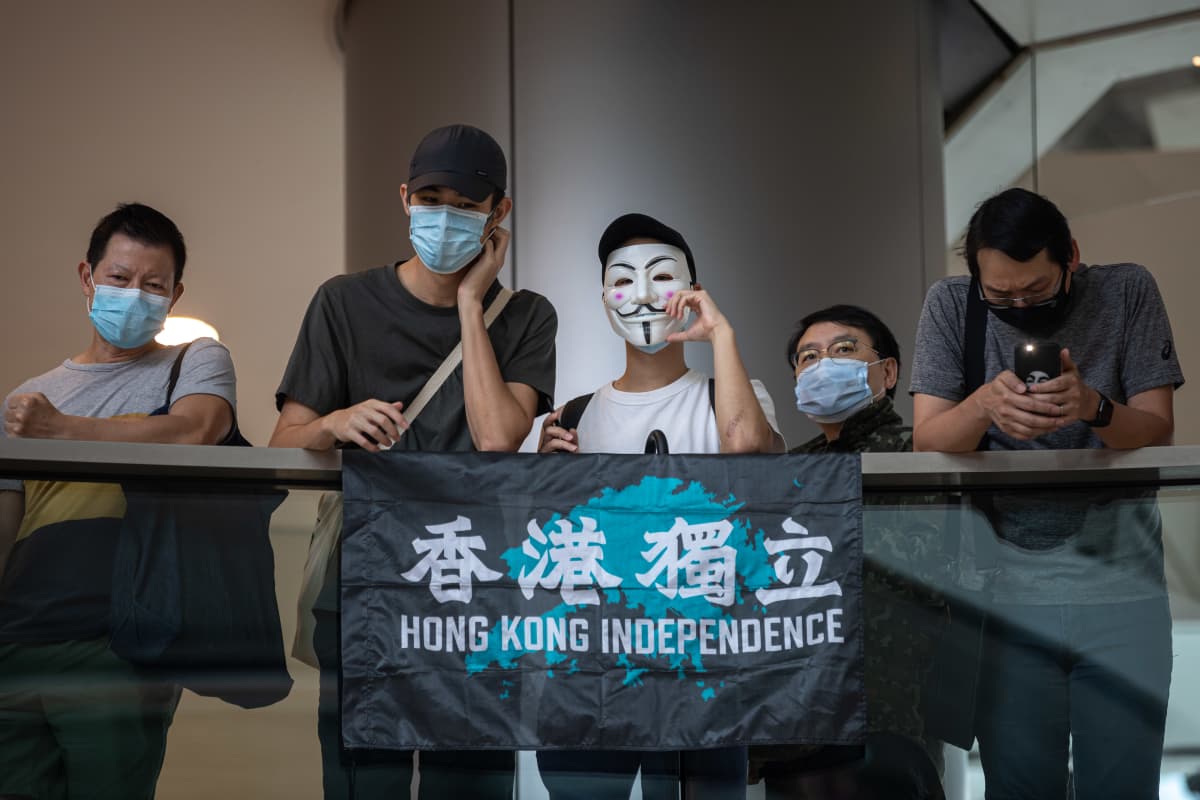 Viisi ihmistä kaiteen takana, osalla kasvoillaan suu-nenäsuoja, yhdellä naamari. Kaiteella kangas, jossa lukee Hong Kong Independence.