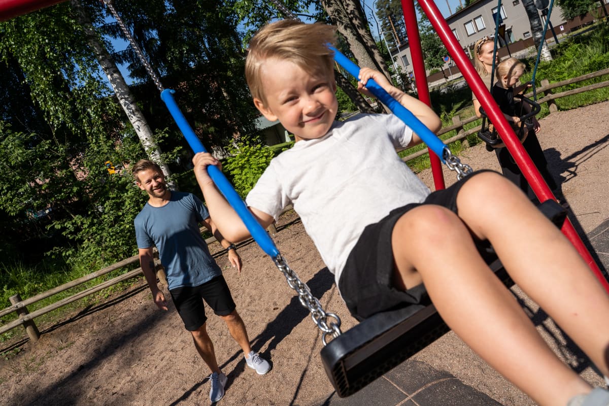Ylhäältä otettu valokuva keinuvasta pojasta, liike pysähtyy lähelle kameraa. Takana isä hymyilee.