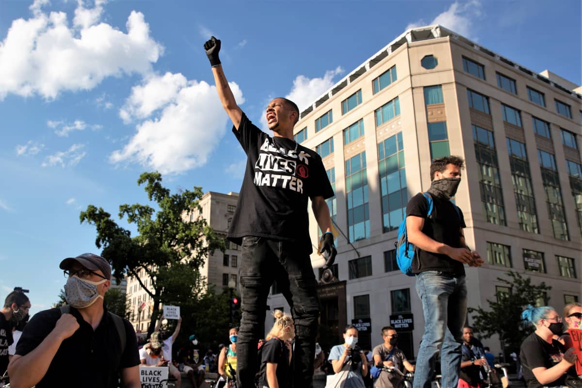 Nuori mies nostaa nyrkkiään ilmaan mielenosoituksessa. Hänellä on yllään musta t-paita. jossa on teksti: Black Lives Matter. Ympärillä on muita mielenosoittajia.