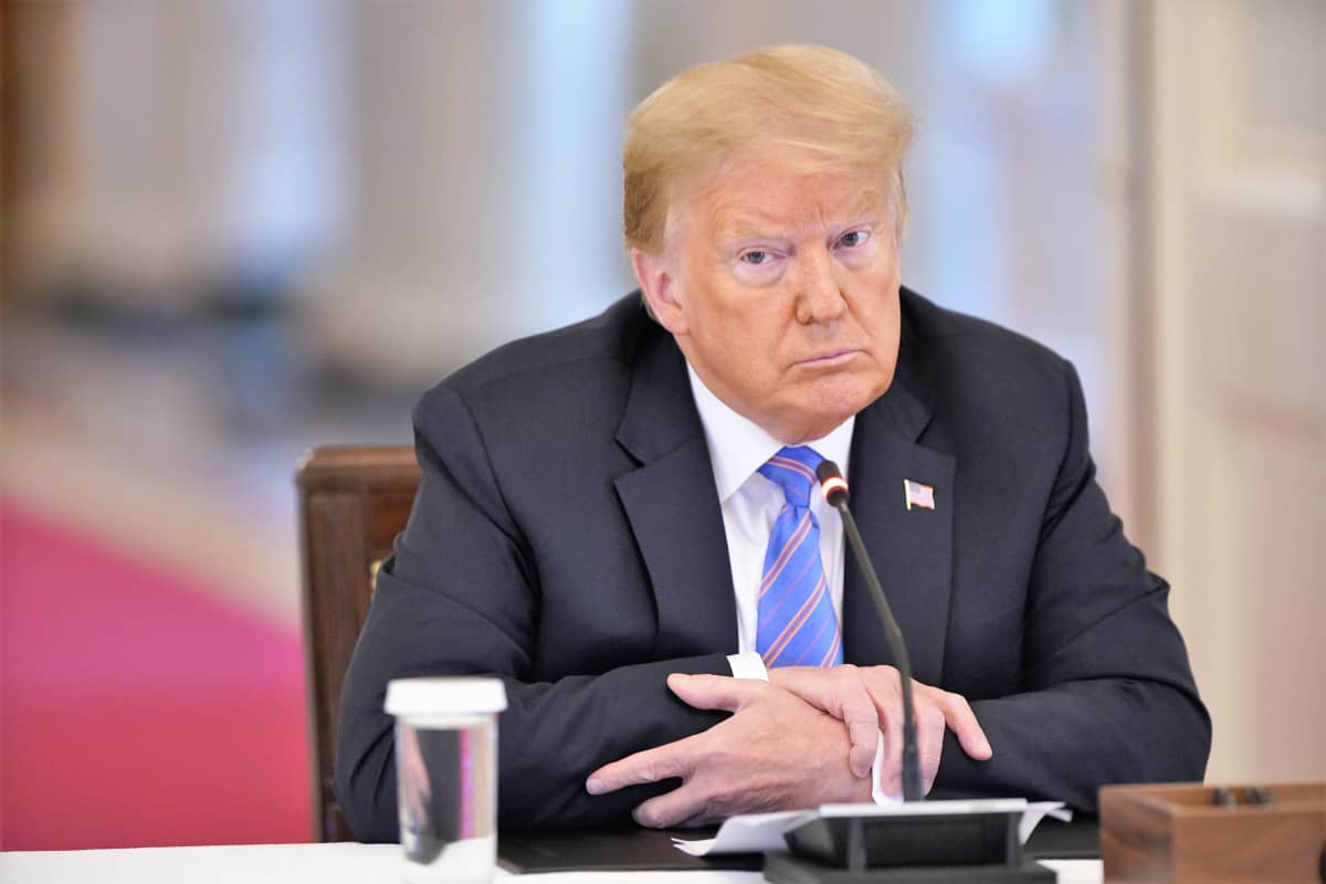 Trump istuu vakavana pöydän takana, nojaten kyynärpäilään pöytään, kädet ristissä. Hänellä on tumma puku, valkoinen kauluspaita, vaaleansininen kravatti ja takin kauluksessa Yhdysvaltain lippua esittävä pinssi.
