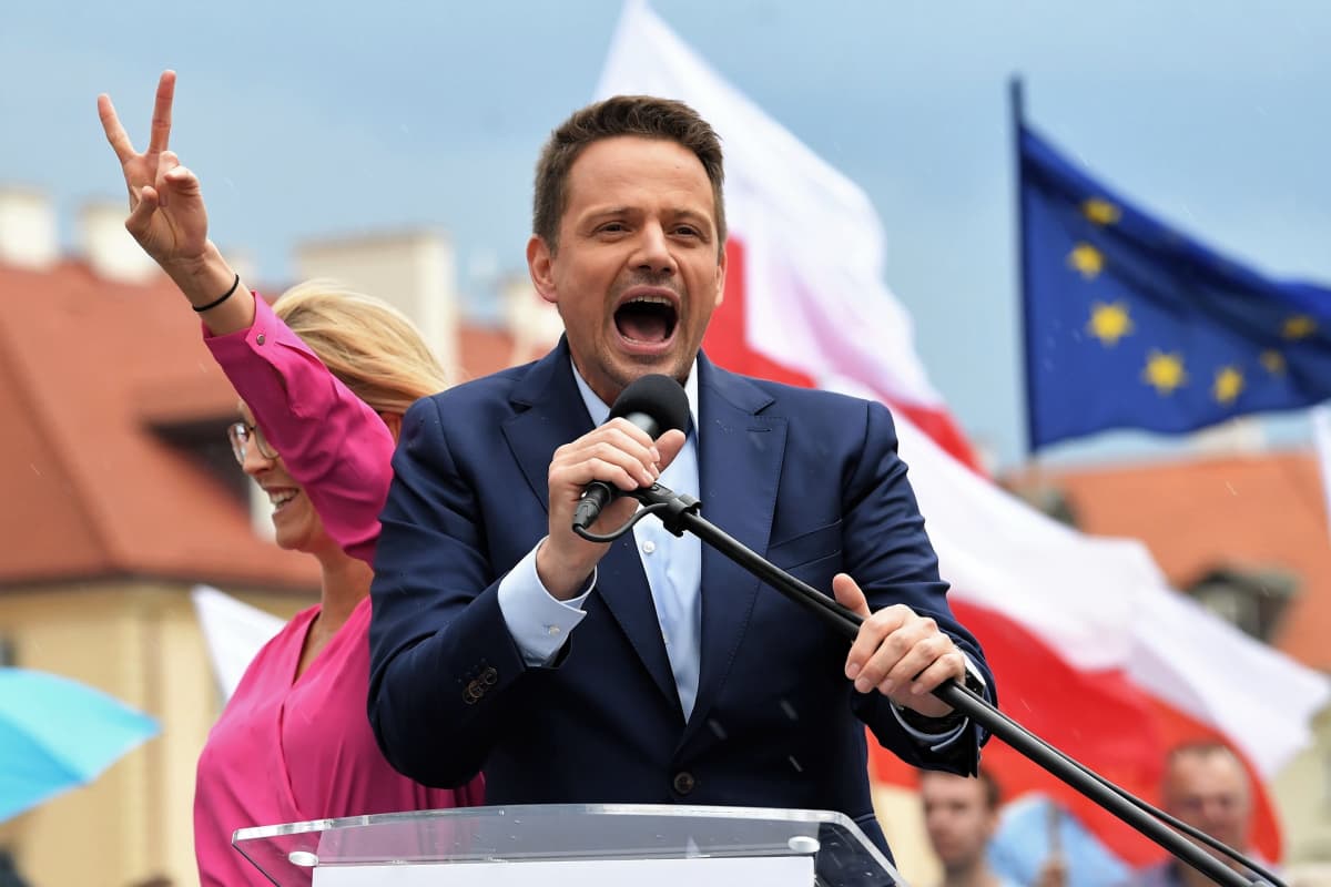 Rafal Trzaskowski huutaa mikrofoniin. Takana seisoo hänen vaimonsa Malgorzata Trzaskowska ja näyttää voitonmerkkiä, Taustalla liehuu EU-lippu. Trzaskowskilla on sininen puku, ei kravattia, hänen vaimollaan on vaaleanpunainen asu.