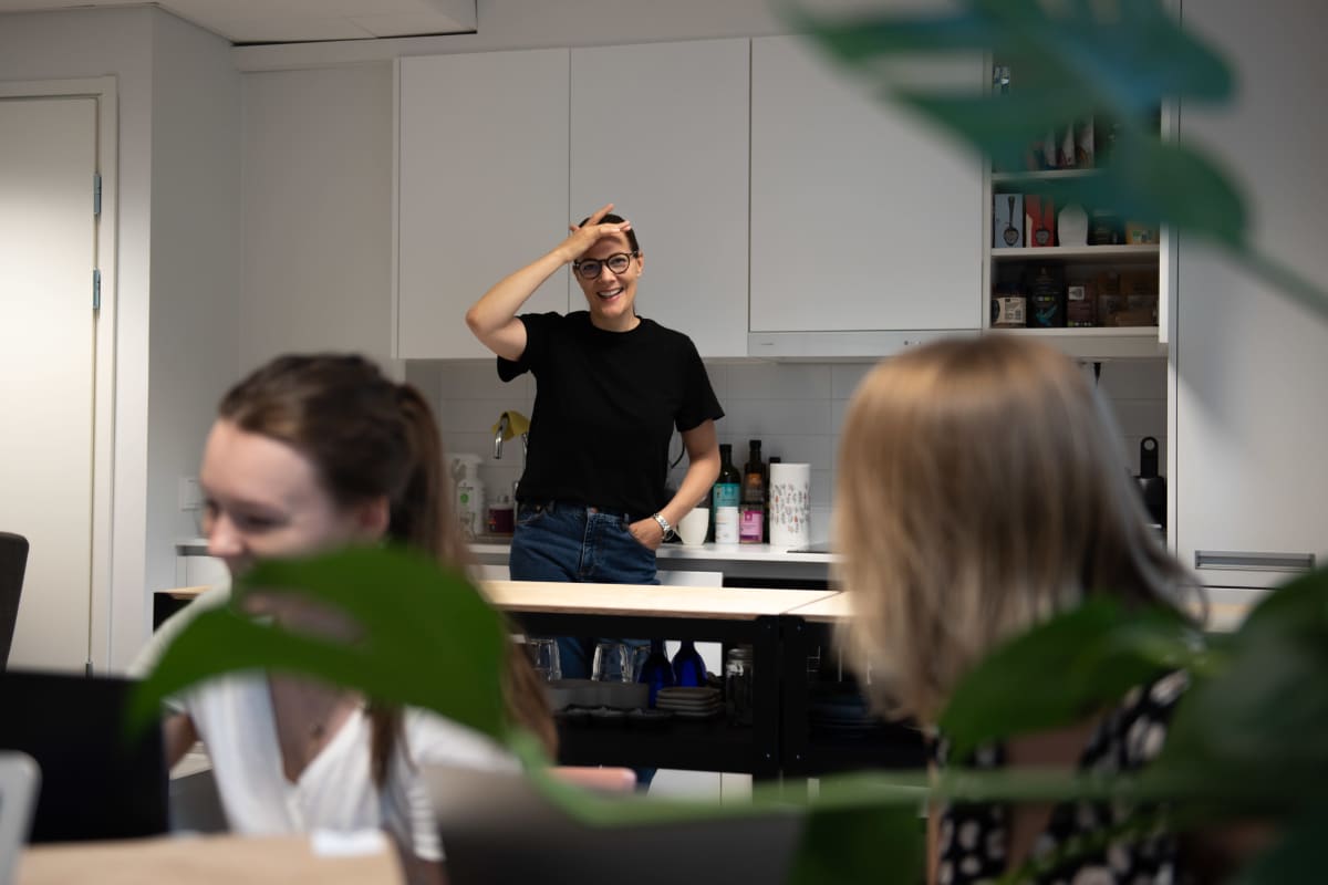Outi Ellilä keittää kahvia toimiston keittiössä, etualalla kesätyöntekijät valmistautuvat kokoukseen.