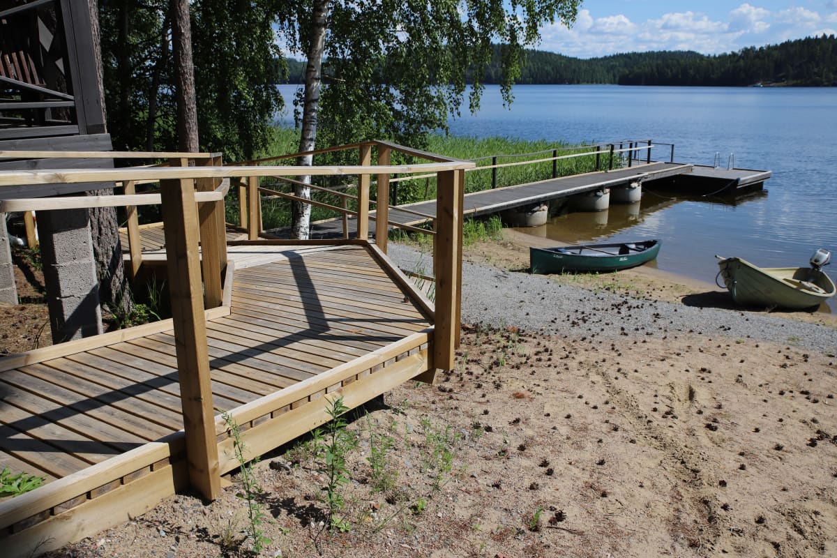Järven rantaan laskeutuu pyörätuoliramppi, joka jatkuu laiturille. Laiturin vieressä hiekkarannalla on kanootti ja soutuvene.