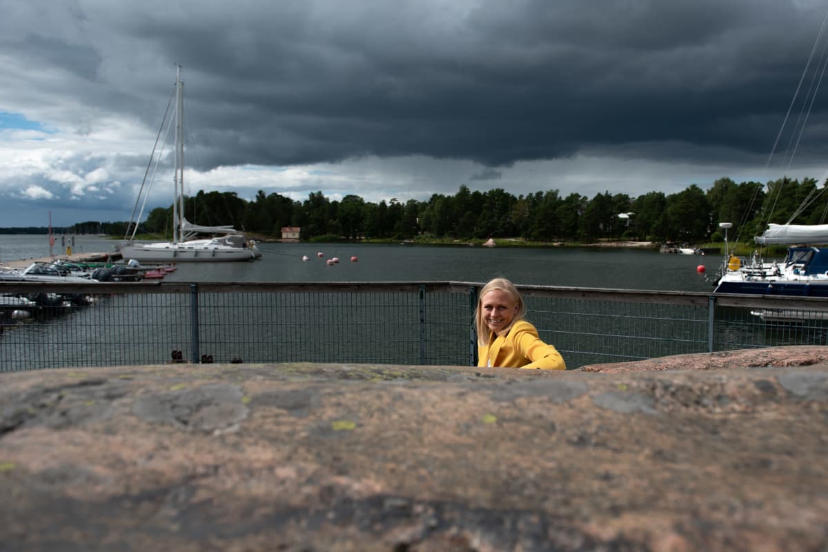 Elina Lepomäki istuu penkillä kallion kupeessa, taustalla näkyy synkkiä sadepilviä.