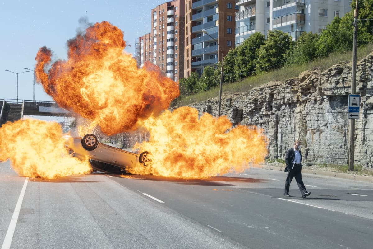 En man står framfölr en bil som exploderat i ett enorms moln av eld.