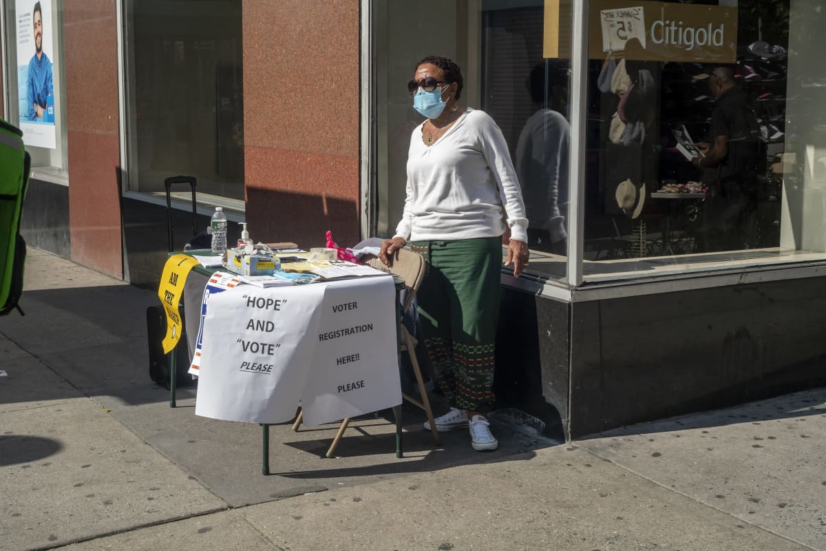 Musta nainen kasvomaskissa pitää äänestäjäksirekisteröitymispistettä New Yorkissa. Nainen seisoo kadunkulmassa pienen pöydän ääressä.