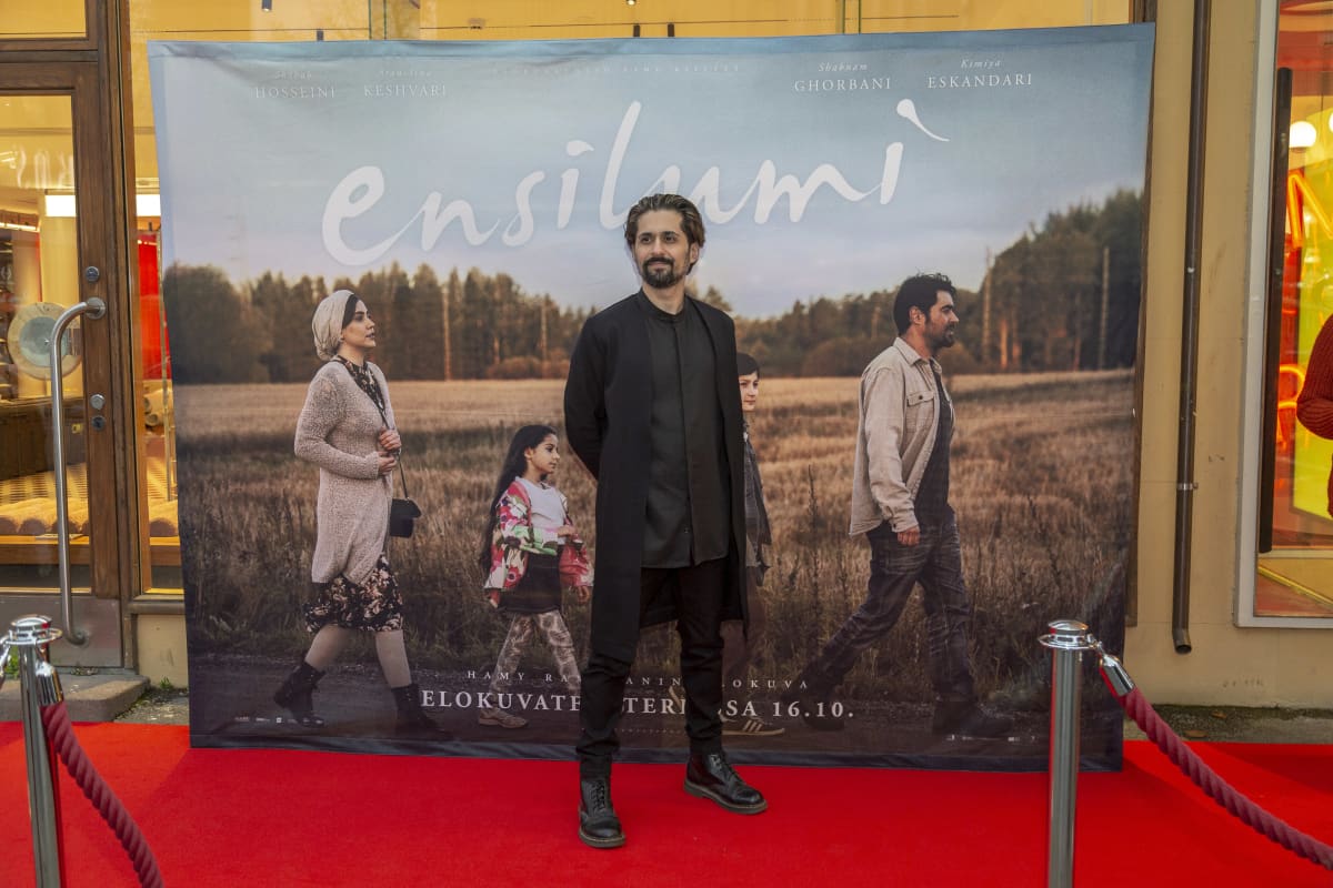 Elokuvaohjaaja Hamy Ramezan poseeraa punaisella matolla Ensilumi-elokuvansa julisteen edessä (kokokuva).