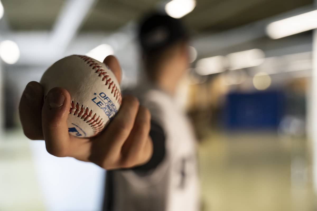 Suomalainen baseball pelaaja Konsta Kurikka heittää baseball palloa käsi taakse ojennettuna. Pallo etualalla juuri ennen heittoa.
