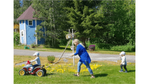 Aikuinen työntää kepillä leikkimönkijää, jonka selässä istuu lapsi. Toinen lapsi seuraa kaksikkoa perässä.