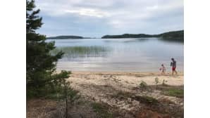 Rantamaisema. Rannalla kävelee lapsi ja aikuinen paljain varpain.